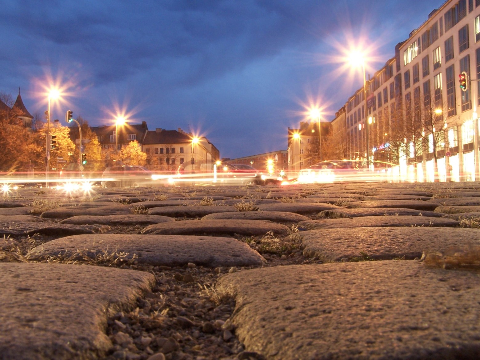 Low-angle view of cobblestone street at dusk with blurred car lights and glowing streetlamps.