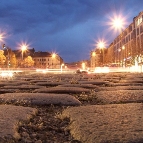 Low-angle view of cobblestone street at dusk with blurred car lights and glowing streetlamps.