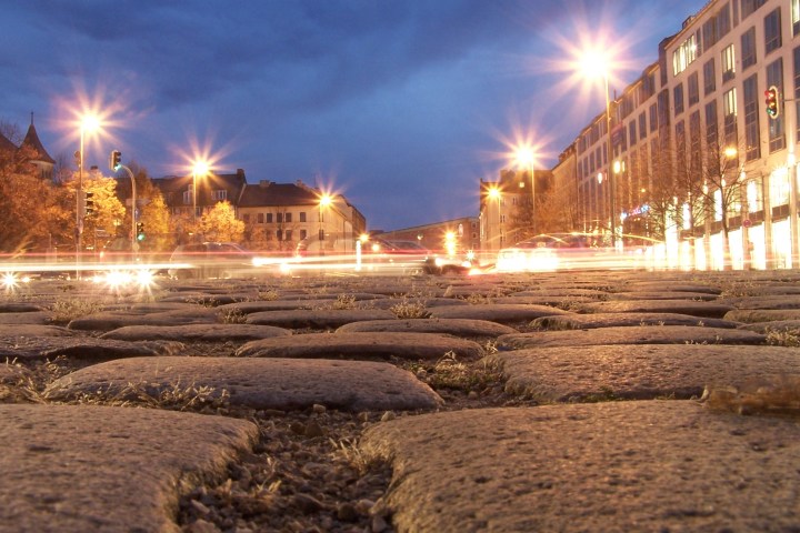 Low-angle view of cobblestone street at dusk with blurred car lights and glowing streetlamps.
