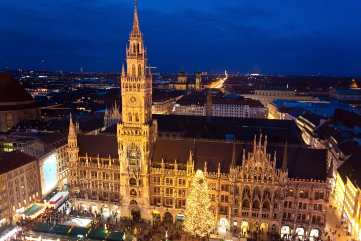 Illuminated building with tall clock tower and Christmas tree in a busy city square at night.
