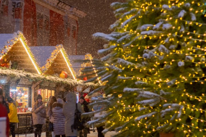 Christmas market with snow, lit huts, and a decorated tree with lights in the foreground.