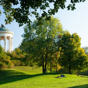 Park landscape with a classical rotunda among trees and people relaxing on grassy hill.