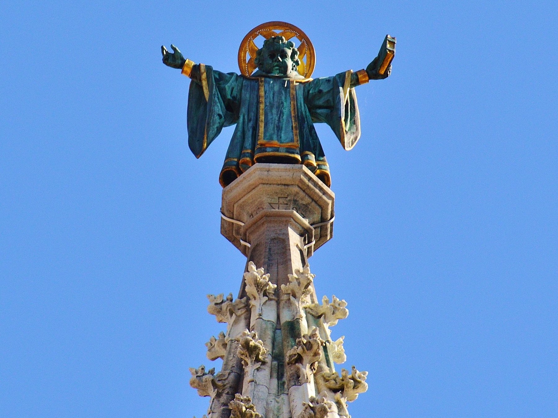Statue of a robed figure with halo atop a decorated spire against a clear blue sky.