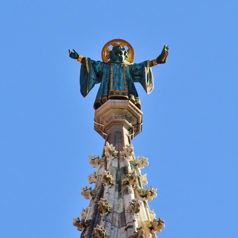 Statue of a robed figure with halo atop a decorated spire against a clear blue sky.