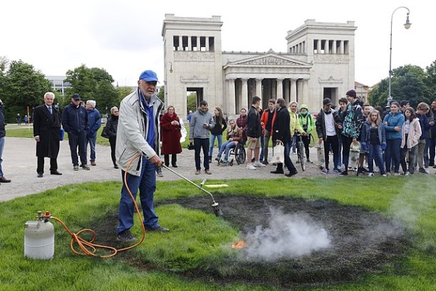 Man with burner torch on grass, crowd watching, memorial for book burnings