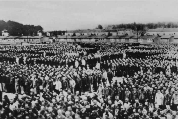 Large group of people in a camp setting with barracks in the background.