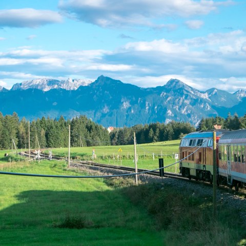 Train on a track with scenic mountain and forest backdrop under a blue, partly cloudy sky.