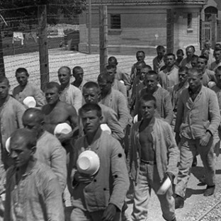 A group of men in uniform walk along a fenced path in a black-and-white photo.