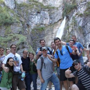 Group of people posing in front of a waterfall and rocky cliff with greenery.