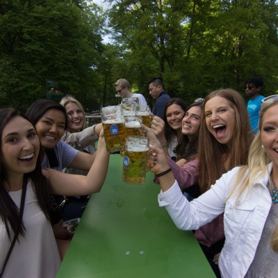 Group of smiling people raising beer mugs at an outdoor picnic table.