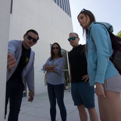 Four people standing outside a building, one pointing at something, wearing casual summer clothing.