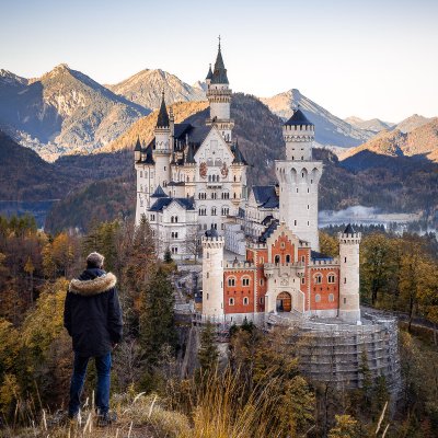 Person in coat overlooking Neuschwanstein Castle with mountains in the background.