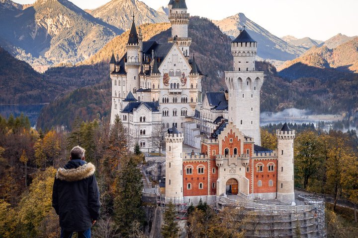 Person in coat overlooking Neuschwanstein Castle with mountains in the background.