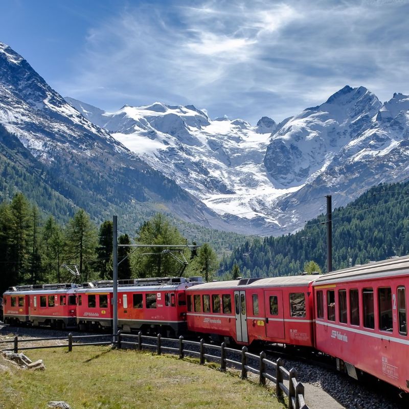 Red train traveling through mountainous landscape with snow-capped peaks and green forests.