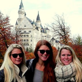 Three women in front of Neuschwanstein Castle with autumn trees in the background.