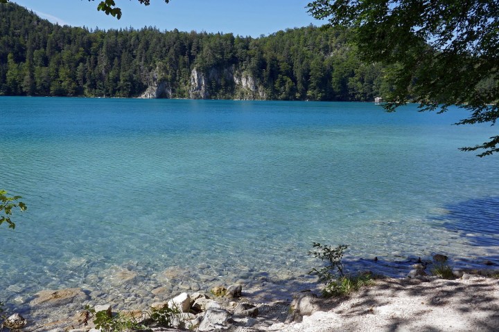 Clear blue lake with sandy shore, surrounded by forested hills under a sunny sky.