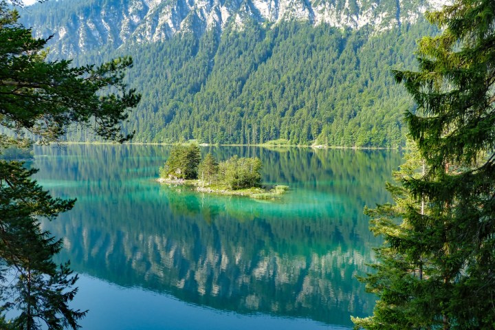 Scenic lake with small island, surrounded by lush green forest and mountains.