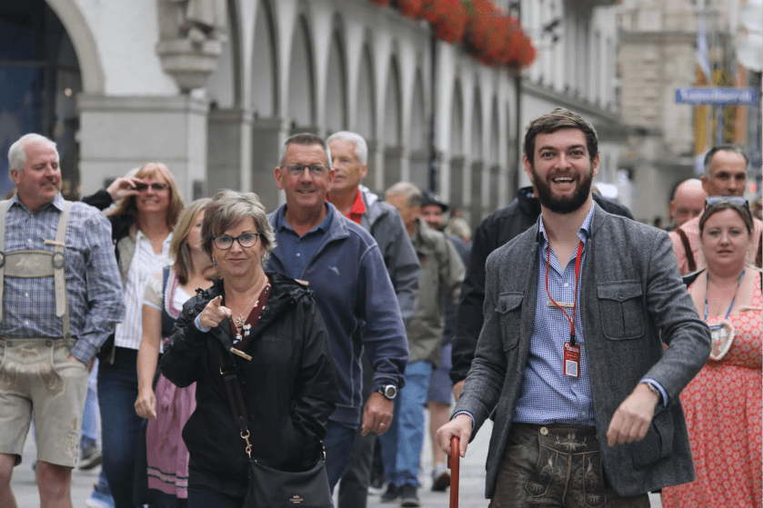 A group of people walking on a street, some smiling, under a row of arches with flowers above.