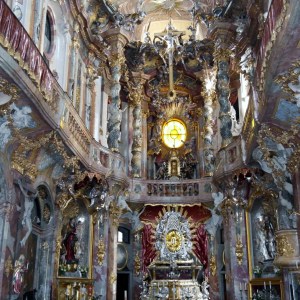Ornate baroque church interior with rich decorations, statues, and a glowing circular window at the altar.