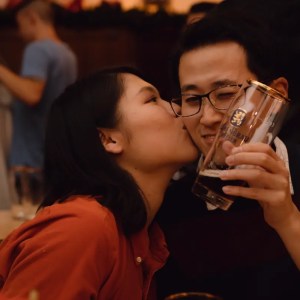 A person kisses another holding a beer glass, both smiling in a cozy, dimly lit setting.