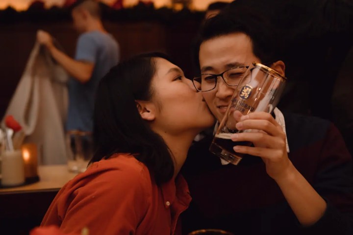 A person kisses another holding a beer glass, both smiling in a cozy, dimly lit setting.