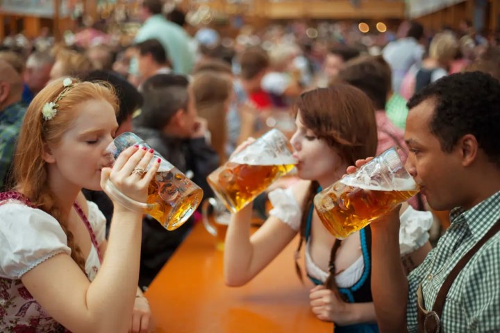 Three people in traditional attire drink large beers at a crowded festival.