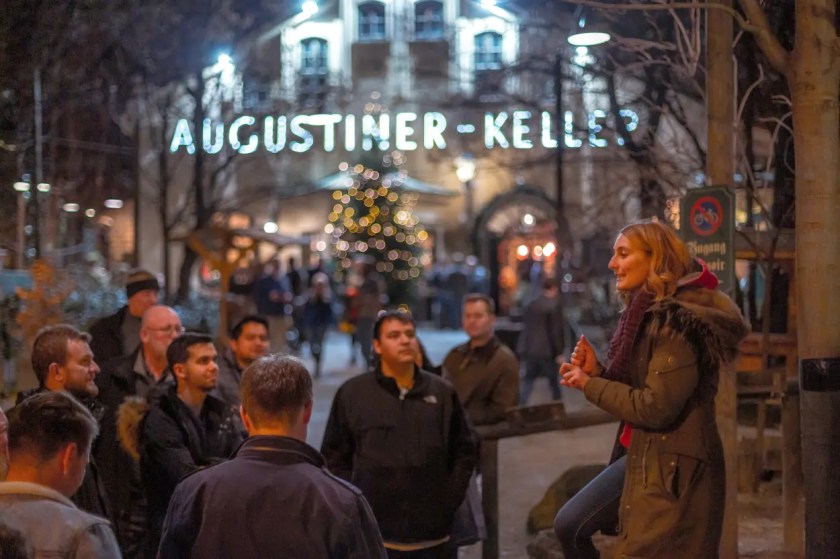 People gathered at a nighttime outdoor event near a building with 'Augustiner-Keller' sign.