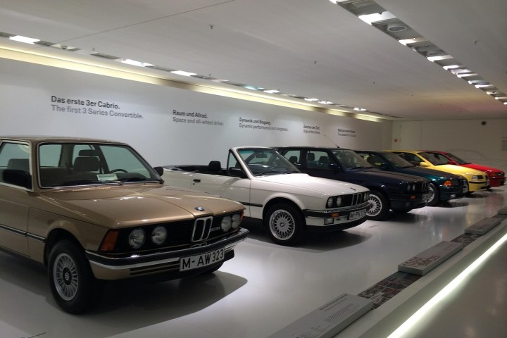 Exhibit of classic BMW 3 Series cars in a showroom with white lighting and descriptive text on walls.