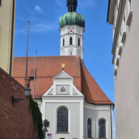 Dachau White church with green onion dome and clock on sloped street.