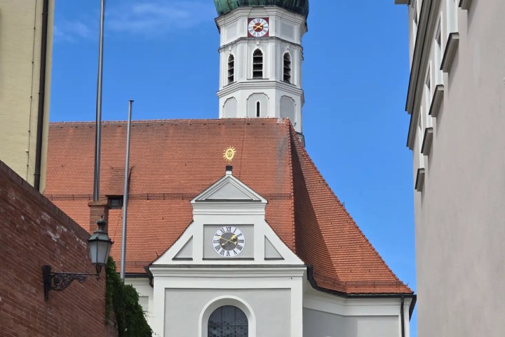 Dachau White church with green onion dome and clock on sloped street.