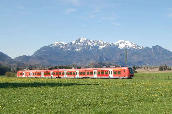 Red train traveling through green field with snow-capped mountains in the background.