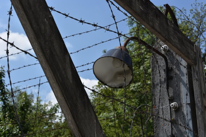 Outdoor light with barbed wire and wooden frame against a blue sky and trees.