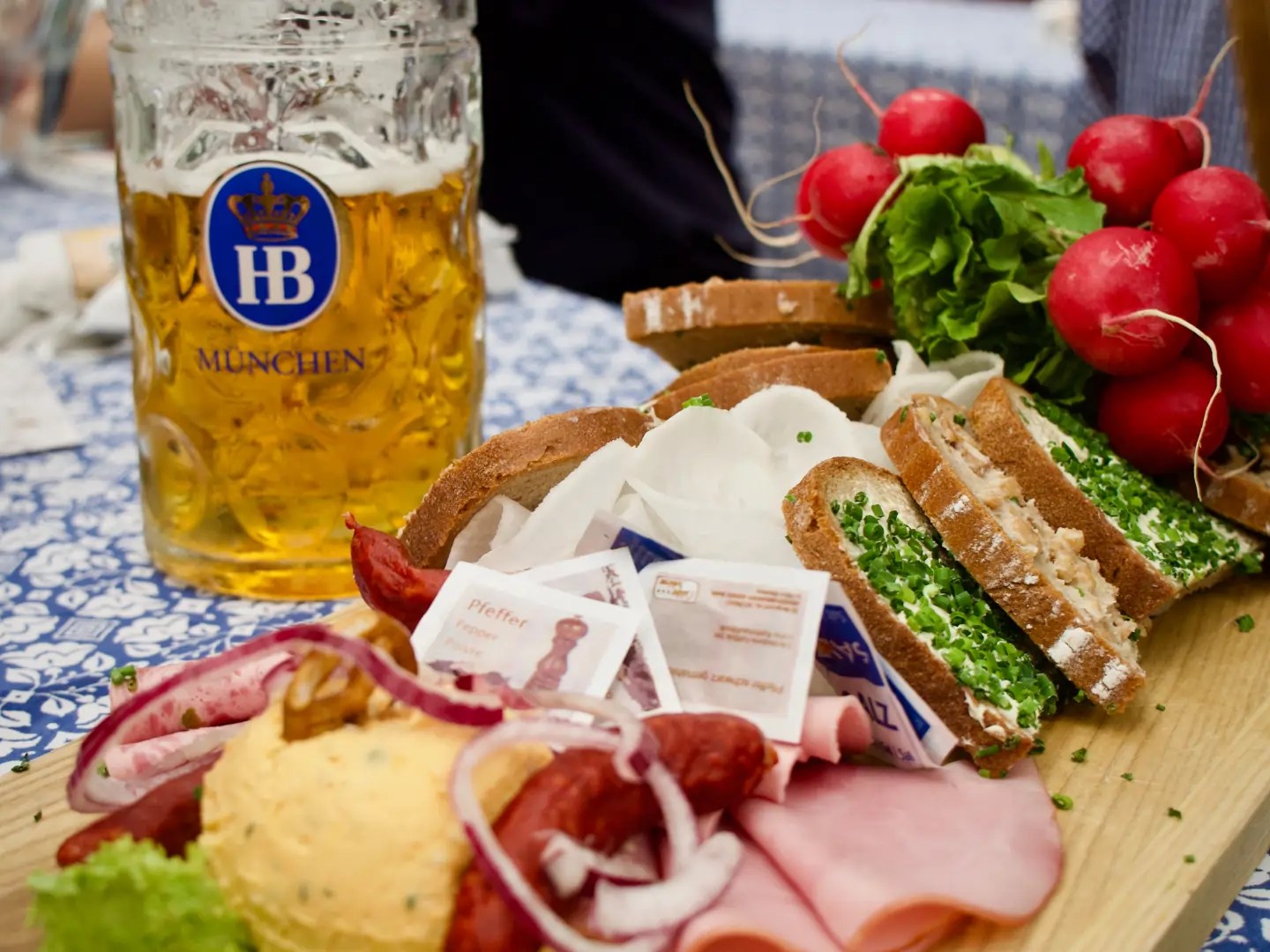 Table with German beer mug, bread, radishes, and sliced meats on a wooden board.