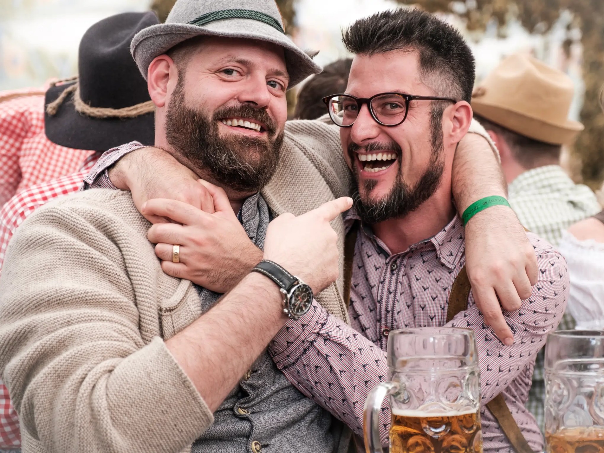 Two bearded men in casual attire smiling and embracing at an outdoor event with beer mugs.