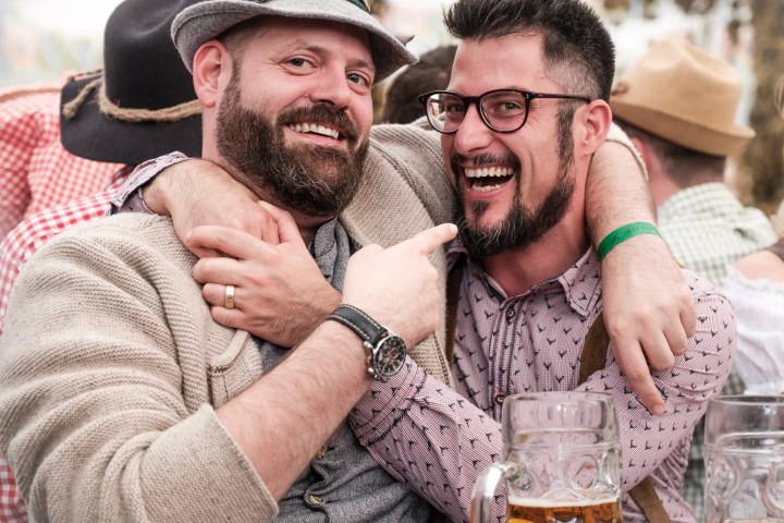 Two bearded men in casual attire smiling and embracing at an outdoor event with beer mugs.