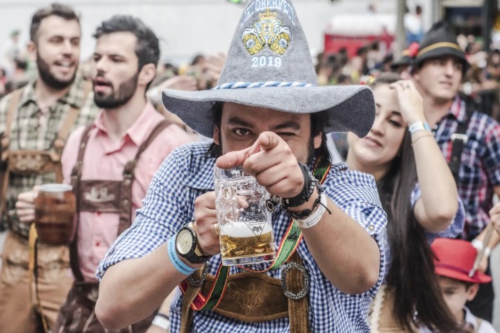 Man in lederhosen and hat holds beer mug, points at camera during festive outdoor event.