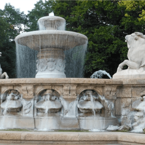 Ornate stone fountain with statues, flowing water, and trees in the background.