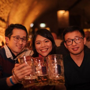 Three friends raising beer mugs in a cozy, warmly-lit bar.