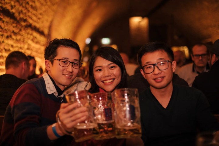 Three friends raising beer mugs in a cozy, warmly-lit bar.