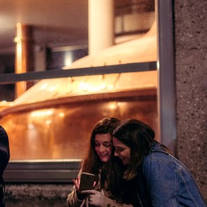Two women smiling at a phone outside a window displaying large brewing equipment.