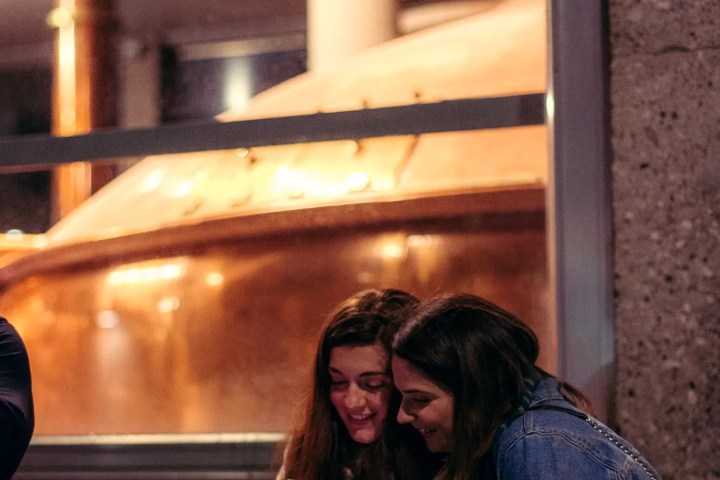 Two women smiling at a phone outside a window displaying large brewing equipment.