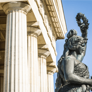 Statue of a woman beside neoclassical columns, holding a wreath, with clear blue sky.