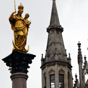 Golden statue on a column next to a Gothic-style tower under a cloudy sky.