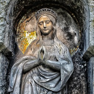 Stone statue of a crowned woman with folded hands in prayer, set in an alcove with mosaic tiles in the background.