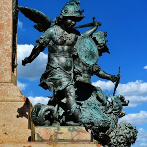 Bronze statue of a winged figure with shield and sword, standing on a creature, against a blue sky.