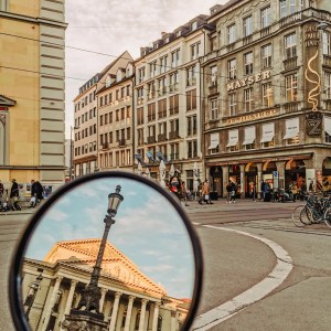 City view with building reflection in a round mirror; people and bikes on street.