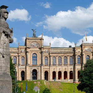 Stone statue in foreground, historic building with arches and flags in the background under a blue sky with clouds.