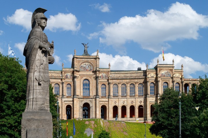 Stone statue in foreground, historic building with arches and flags in the background under a blue sky with clouds.