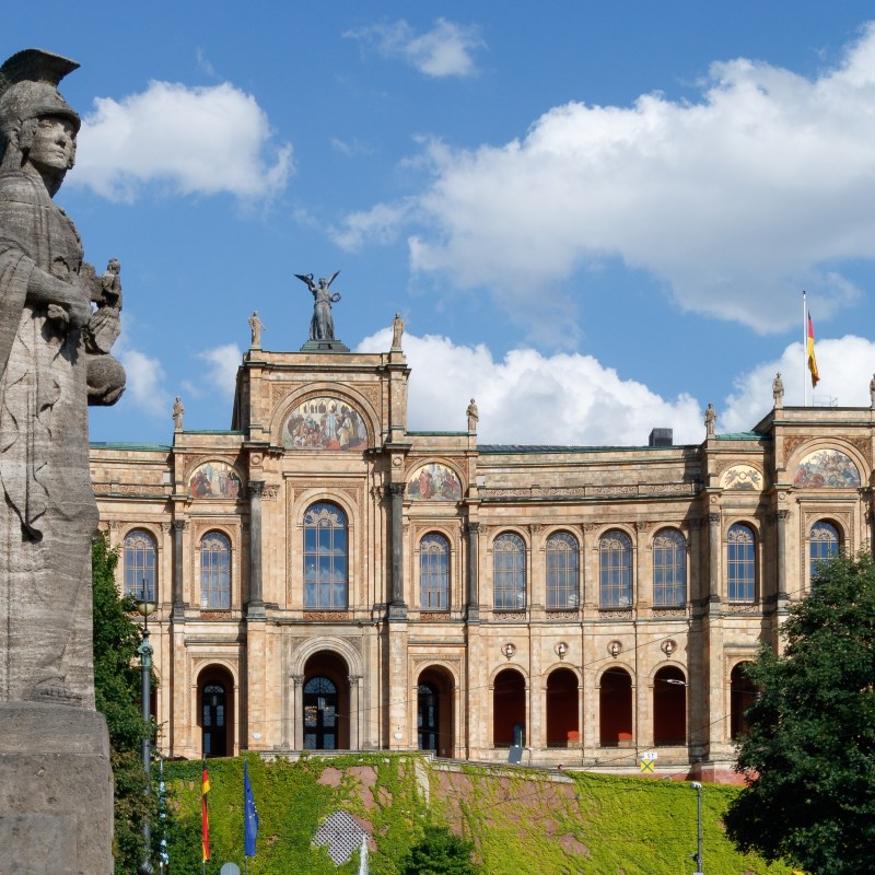 Stone statue in foreground, historic building with arches and flags in the background under a blue sky with clouds.