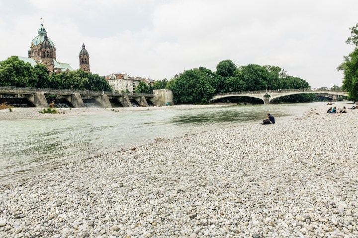 Riverbank with pebbles, bridge, trees, and a church in the background, people sitting on the shore.